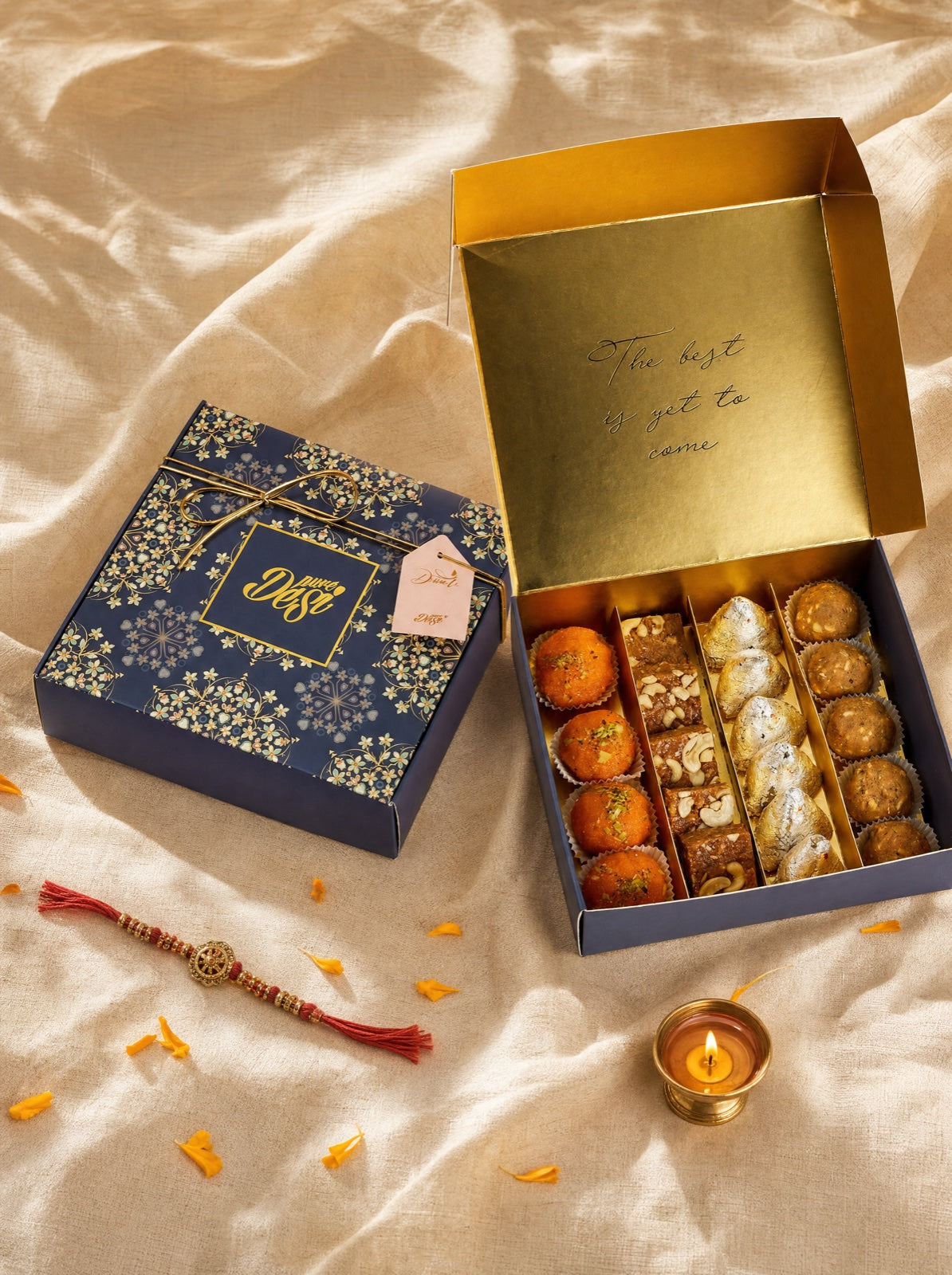 A Pure Desi build-your-own gift box open on cream cloth beside a rakhi thread and a lit diya