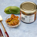 Pure Desi mango pickle in a bowl with two green mangoes and the 300g jar